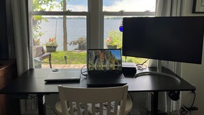 A room with a desk, a laptop, and a monitor. There is a window with a view of a body of water and greenery outside.