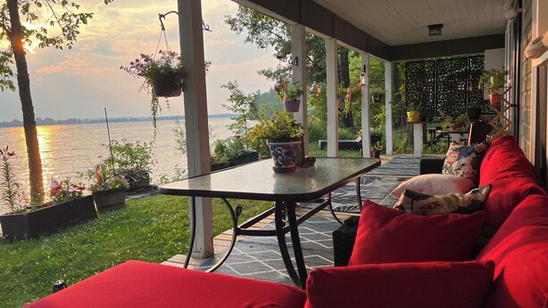 A covered outdoor seating area with a view of a body of water, a table with a hanging plant, and red cushions.