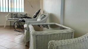 Una habitación con un sofá y sillones de mimbre blanco, una mesa con cubierta de vidrio y vistas a través de una ventana con barrotes.