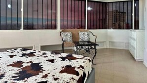 Una habitación con una cama, una silla, un televisor y ventanas grandes con barrotes.