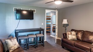 Ein Wohnzimmer mit einem braunen Ledersofa, einem dunklen Holztisch und zwei Stühlen, einem an der Wand befestigten Flachbildfernseher, einem Deckenventilator und einem Durchgang zu einem Schlafzimmer mit Etagenbett.