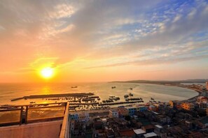 Un paisaje urbano costero con un atardecer sobre el mar, edificios y un puerto deportivo.
