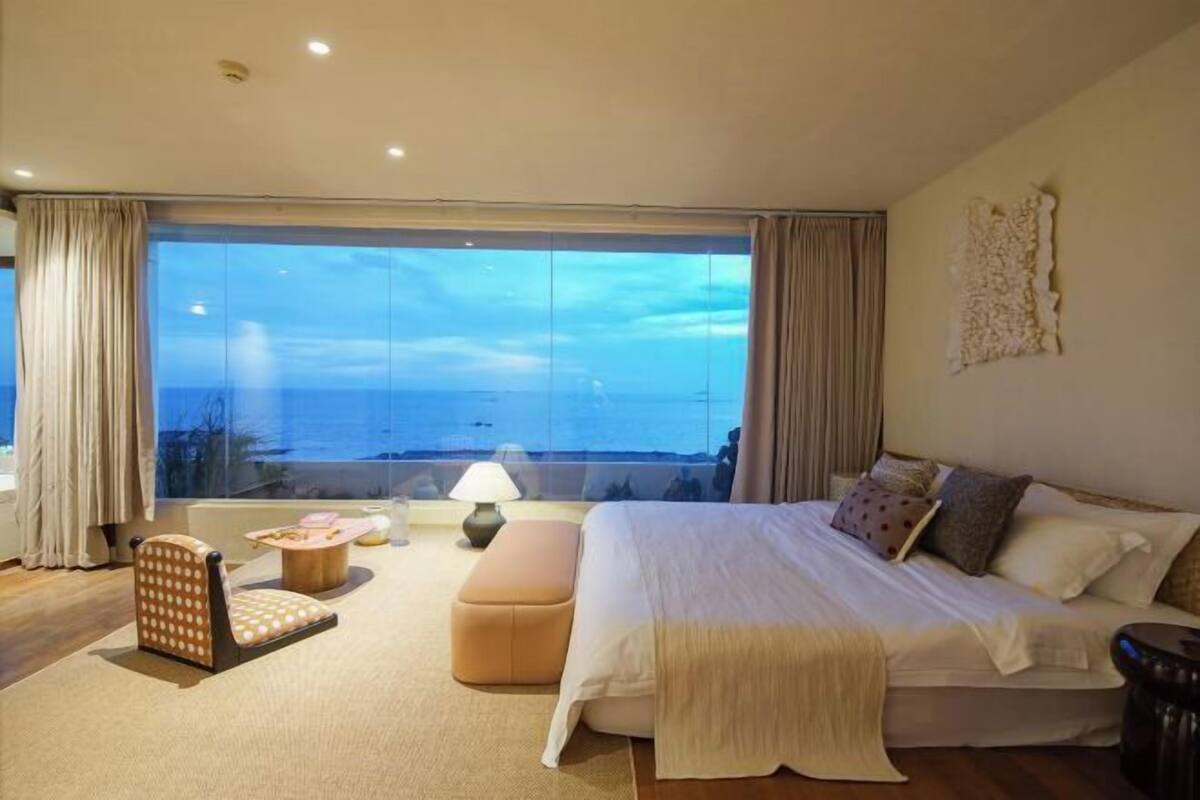 Ein Schlafzimmer mit einem großen Bett, einem Sessel, einem kleinen Tisch und Blick aufs Meer.