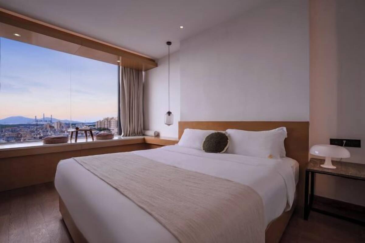 Ein Hotelzimmer mit einem großen Bett, einem Nachttisch, einer Lampe, Blick auf die Stadt und einem Vorhangfenster.