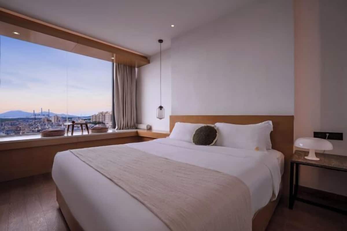 A hotel room with a large bed, a bedside table, a lamp, a view of the city, and a window with curtains.
