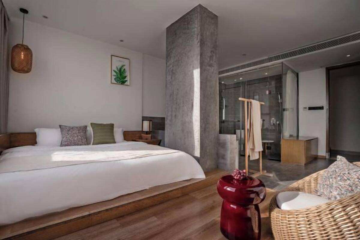 Ein modernes Hotelzimmer mit einem großen Bett, einer roten Vase, einem Korbstuhl und einer Duschkabine aus Glas.