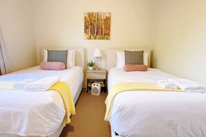 Ein Hotelzimmer mit zwei Betten, jeweils mit weißer Bettwäsche und einer gelben Tagesdecke, einem kleinen Nachttisch mit einer Lampe sowie einem an der Wand hängenden Gemälde.