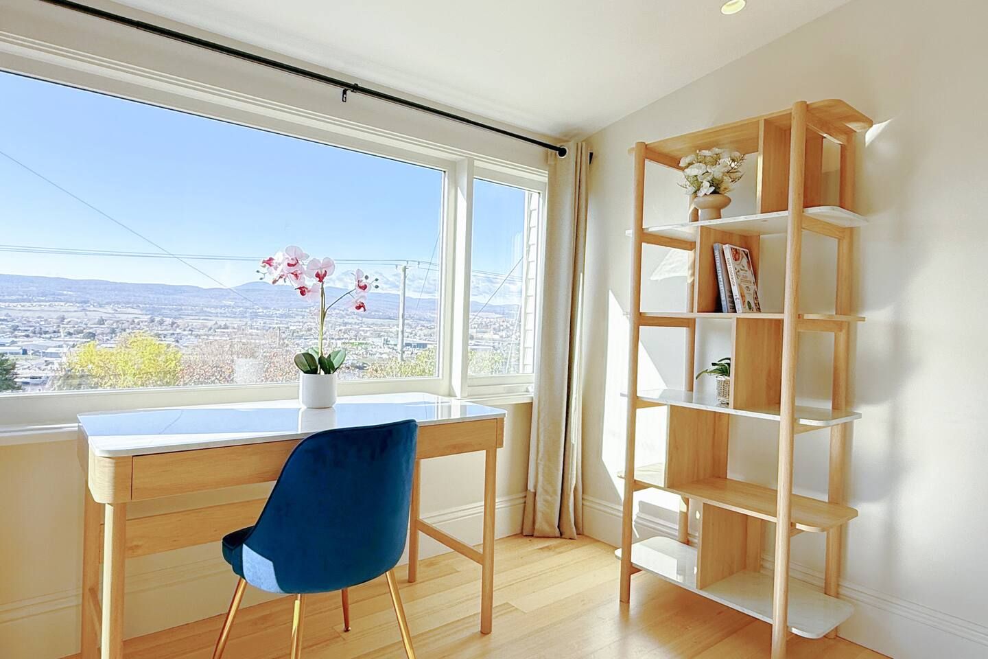 A room with a desk, a blue chair, a wooden bookshelf, and a window with a city view.