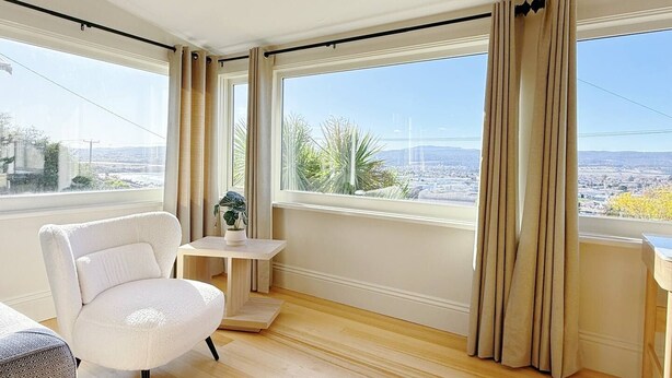 A modern room with large windows, a white chair, a small table with a plant, and a view of a cityscape.