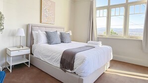 A bedroom with a large bed, a bedside table with a lamp, a framed artwork on the wall, and a window with a view.