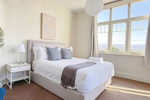 A bedroom with a large bed, a bedside table with a lamp, a framed artwork on the wall, and a window with a view. - StayAU 2BR Home w/ Stunning Views in Tasmania (Trevallyn)