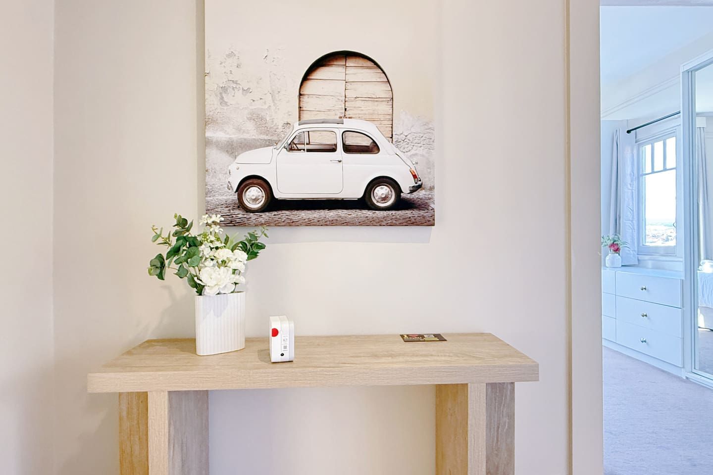 Una habitación con un cuadro colgado en la pared que muestra un coche, una mesita con un jarrón blanco y un control remoto, y una ventana con vistas.