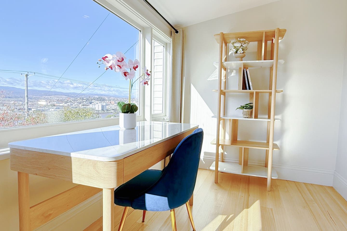 Ein modernes Zimmer mit Schreibtisch, blauem Sessel, Holzregal und Blick auf eine Stadtlandschaft durch große Fenster.