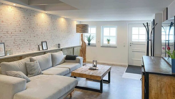 A modern living room with a white brick wall, a sectional sofa, a wooden coffee table, and a minimalist decor style. - Lüttjet Huus (Lügde)