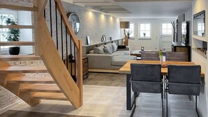 A modern living room with a staircase, a dining area with a wooden table and chairs, and a gray sofa. - Lüttjet Huus (Lügde)