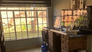 Una cocina con estufa a gas, un cilindro de gas azul, un cubo de basura y una ventana con persianas.