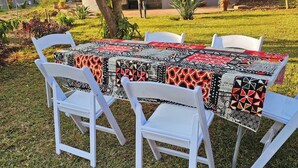 Un área de comedor al aire libre con una mesa cubierta con un mantel estampado, rodeada de sillas blancas, ubicada sobre un césped con una casa y árboles al fondo.