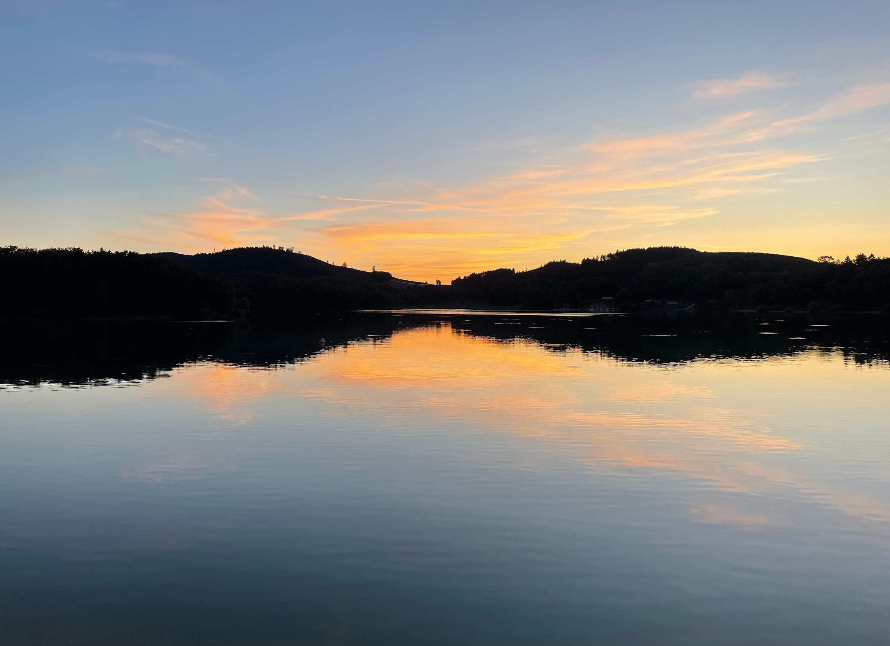 Un lac paisible aux eaux claires, dont le reflet se mire dans les collines environnantes, couvertes de forêts.