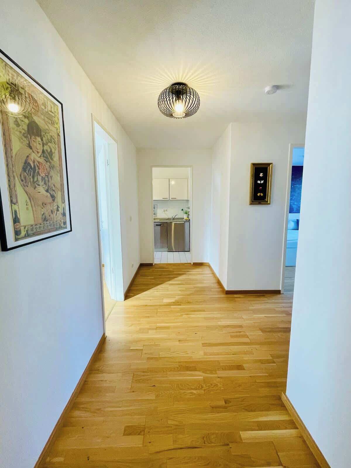 A hallway with wooden flooring, white walls, a ceiling light fixture, and a kitchen area visible through an open door.