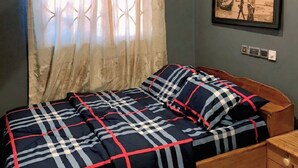 A bedroom with a bed, a nightstand, a framed picture on the wall, and a window with curtains. - StayComidad
Your comfort, Our Priority. (Tamale)