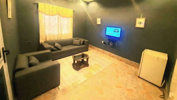 A living room with a sectional sofa, a coffee table, a flat-screen TV, and a refrigerator. - StayComidad
Your comfort, Our Priority. (Tamale)