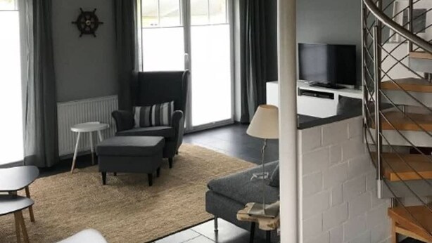 Een moderne woonkamer met een bank, een poef en een televisie op een dressoir. Er is een trap met een metalen leuning die naar een verdieping leidt.