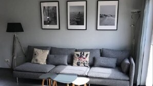 Een moderne woonkamer met een grijze bank, een klein bijzettafeltje en een zwart voetenbankje. Er hangen drie ingelijste foto’s aan de muur en links staat een vloerlamp.