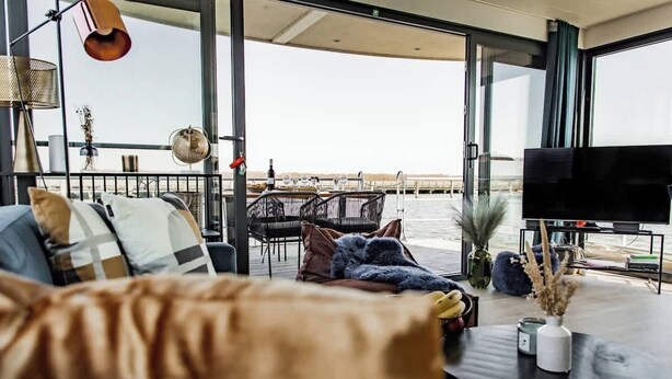 A modern living room with a sofa, a coffee table, and a TV. There is a balcony with a view of the water. - Hausboot Küstennebel 2 (Ribnitz-Damgarten)