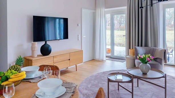 Een moderne woonkamer met een flatscreen-tv, houten meubels, een eettafel gedekt met borden en glazen, en uitzicht op de omgeving.