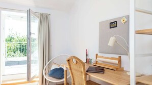 Una habitación con escritorio de madera, una silla de mimbre, una silla blanca con cojín azul, una lámpara y vistas a zonas verdes a través de una puerta corredera.