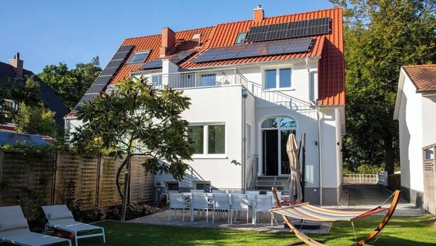 Una casa moderna de dos pisos con techo de tejas rojas, paneles solares y un pequeño jardín con sillas blancas y una hamaca.