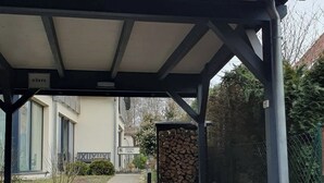 Un acceso cubierto con un camino de ladrillo que lleva a un edificio, una pila de leña y una valla metálica.