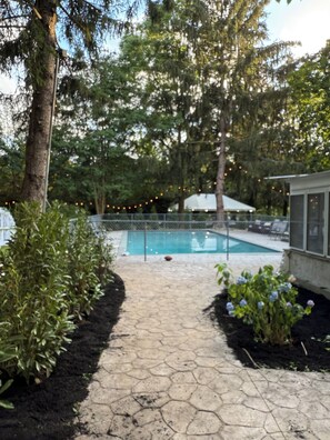 A swimming pool with a hexagonal tile pathway leading to it, surrounded by trees and a fenced area with lounge chairs.