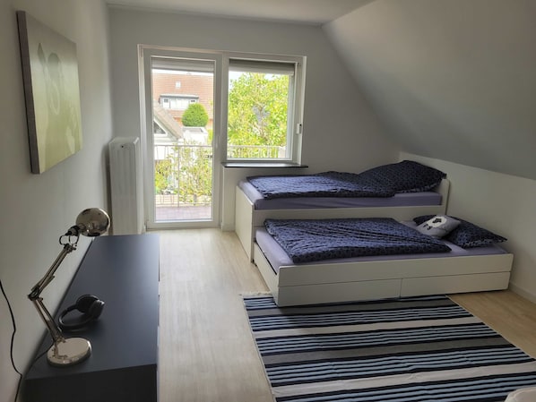 A bedroom with two beds, a desk, a chair, a computer, and a window with a view of a garden.