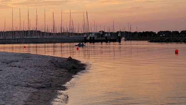 Ein ruhiges Ufer mit anlegenden Booten, einem Sandstrand und einer Pier, die sich ins Wasser erstreckt.