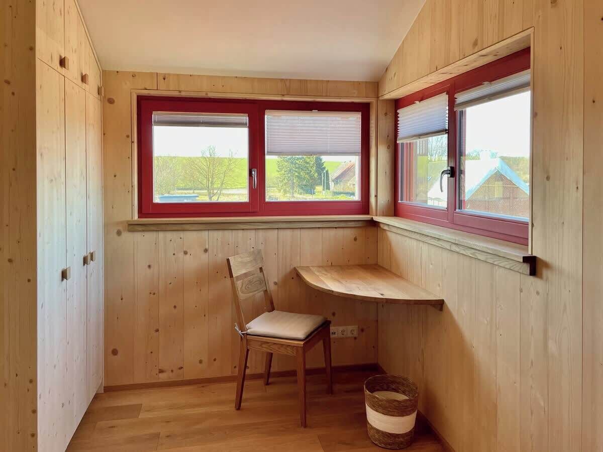 Una habitación pequeña con paredes de madera y una ventana con marco rojo. Una silla de madera está apoyada contra la pared, y una pequeña mesa está colocada debajo de la ventana.