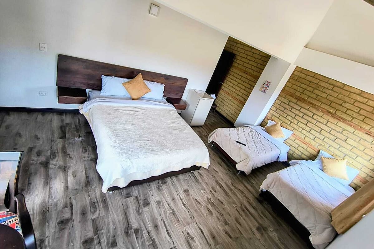 Une chambre d’hôtel avec un grand lit, deux petits lits, un plancher en bois, un mur en briques et un plafond blanc.
