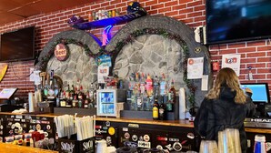 Een bar met een bakstenen muur, een televisie boven de bar en een toonbank met verschillende flessen en een neonbord.