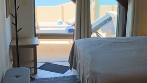 A bedroom with a bed, a bench, a small table, and a view of the sea through a window with curtains. - Seaview Tranquility Steps to Ocean's Edge, Sunsets (Rethimno)