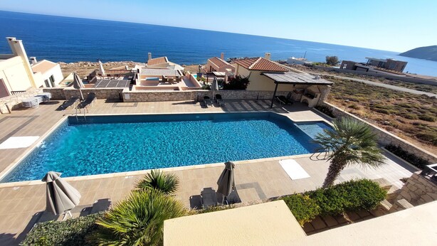 A swimming pool with lounge chairs and umbrellas, surrounded by buildings with a view of the sea. - Seaview Tranquility Steps to Ocean's Edge, Sunsets (Rethimno)