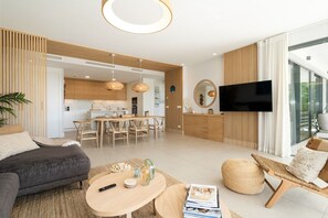 A modern living room with a sofa, coffee table, and a TV. There's a dining area with a table and chairs, and a kitchen in the background. - Aqualina de Madeira (Benahavís)