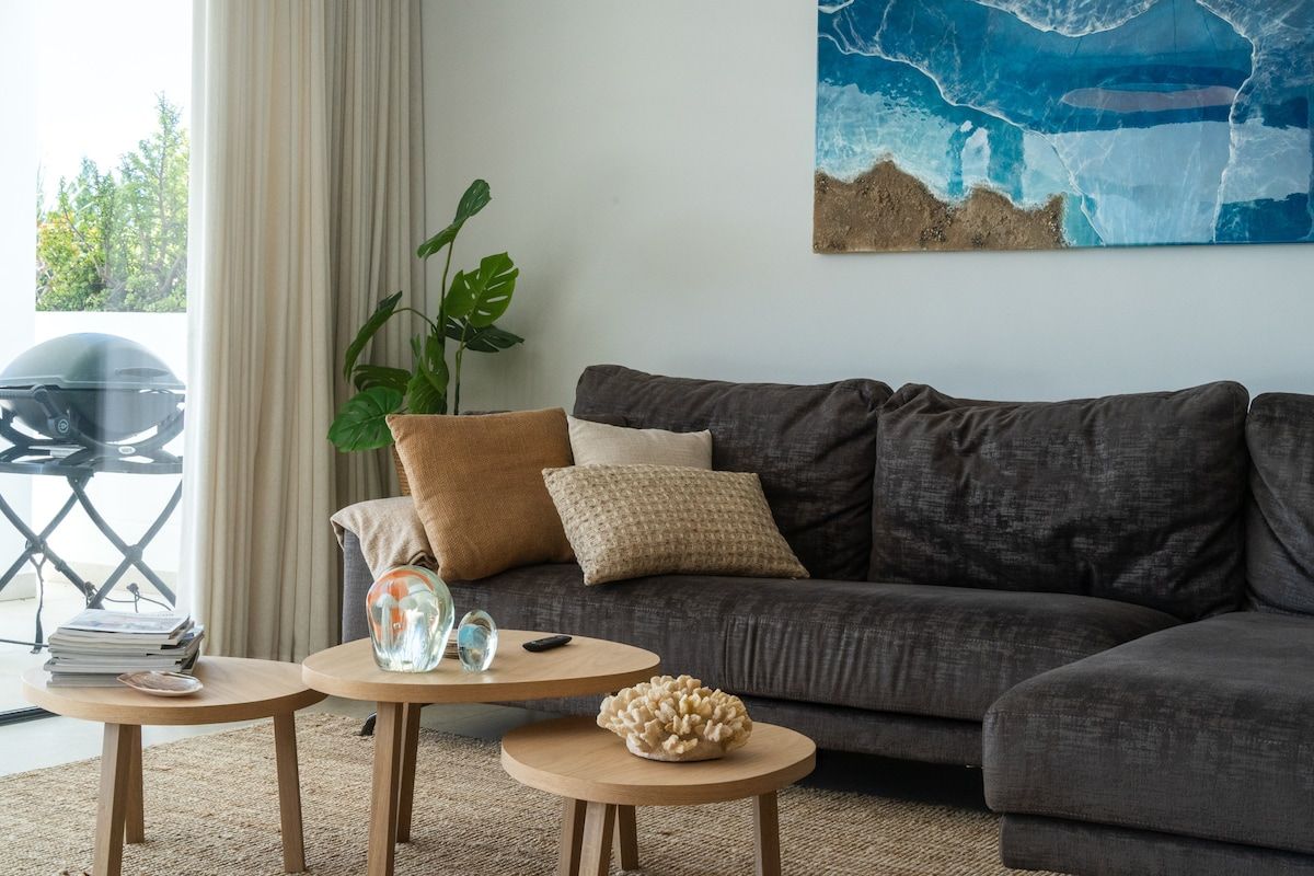 En moderne stue med en mørkegrå sofa, to små træborde, en potteplante og et stort abstrakt maleri på væggen.