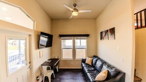 Un espace de vie compact doté d’un plancher en bois foncé, d’un canapé en cuir noir, d’une table à manger avec des chaises, d’un ventilateur de plafond et d’un téléviseur fixé au mur.