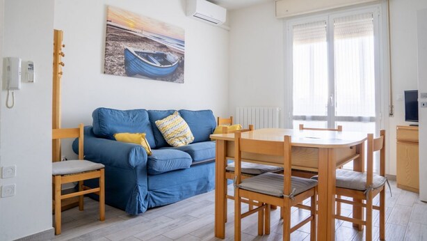 Een woonkamer met een blauwe bank, een houten eettafel en stoelen, een schilderij aan de muur en een raam met jaloezieën.