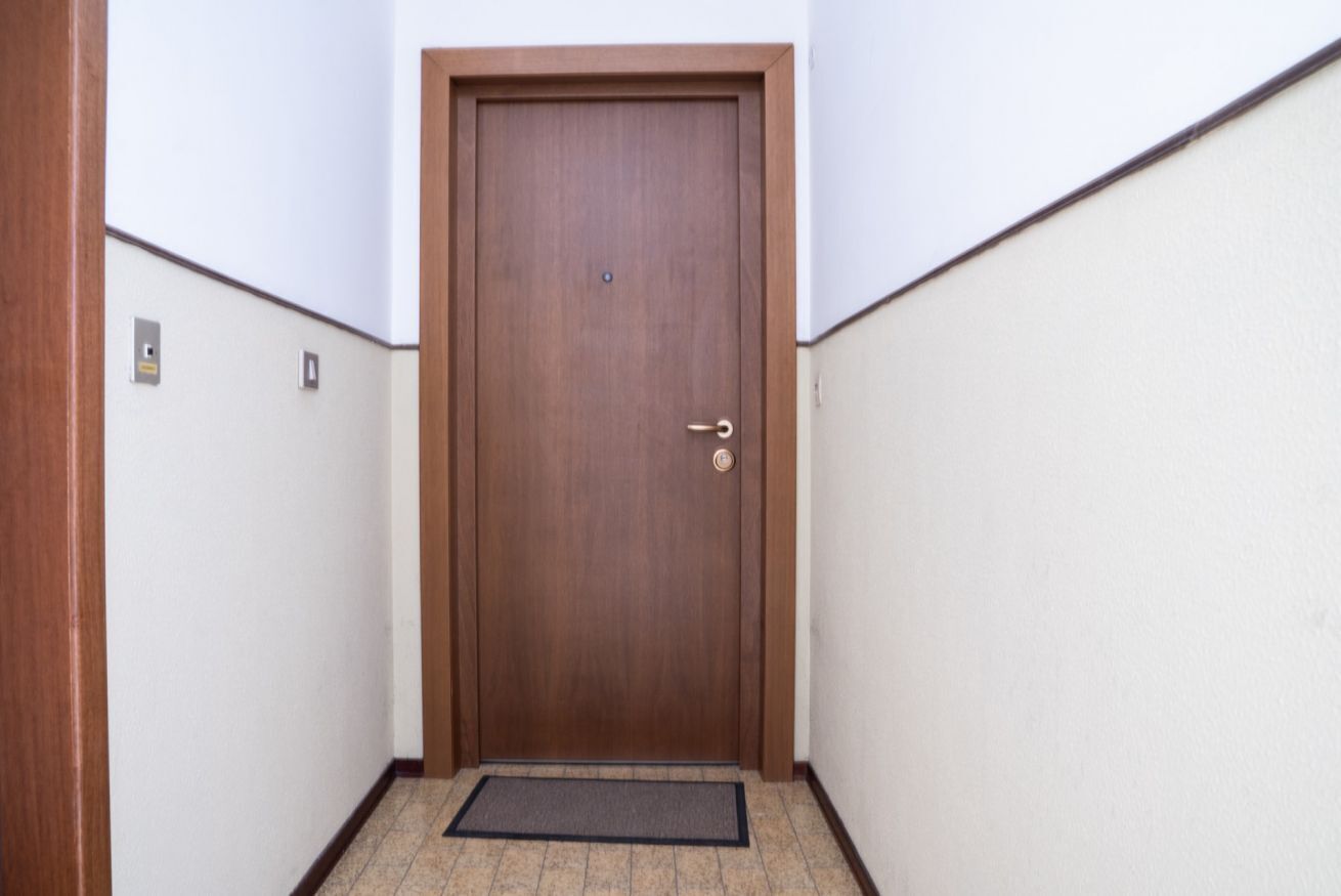 A wooden door in a hallway with a doormat and a light switch on the wall.