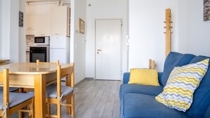 Een knus leefgedeelte met een eethoek, een kitchenette, een bank met opvallende kussens en een gitaar die tegen de muur staat.