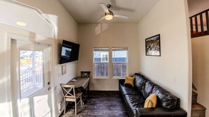 A compact living space with a ceiling fan, a TV mounted on the wall, a black leather couch, a wooden table, and a window with blinds. - Moab Tiny Home Near Arches | Sleeps 6 | King + Loft | Mill Creek Falls (Moab)