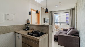 A modern kitchen with a gas stove, a built-in oven, and a small refrigerator. There is a dining area with a sofa and a table, and a balcony with a view of the city. - Apartment Next to Flamboyant Shopping Mall - ESS (Goiânia)