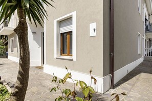 A building with a stone-paved courtyard, a window with a wooden shutter, and a small white sign on the wall.