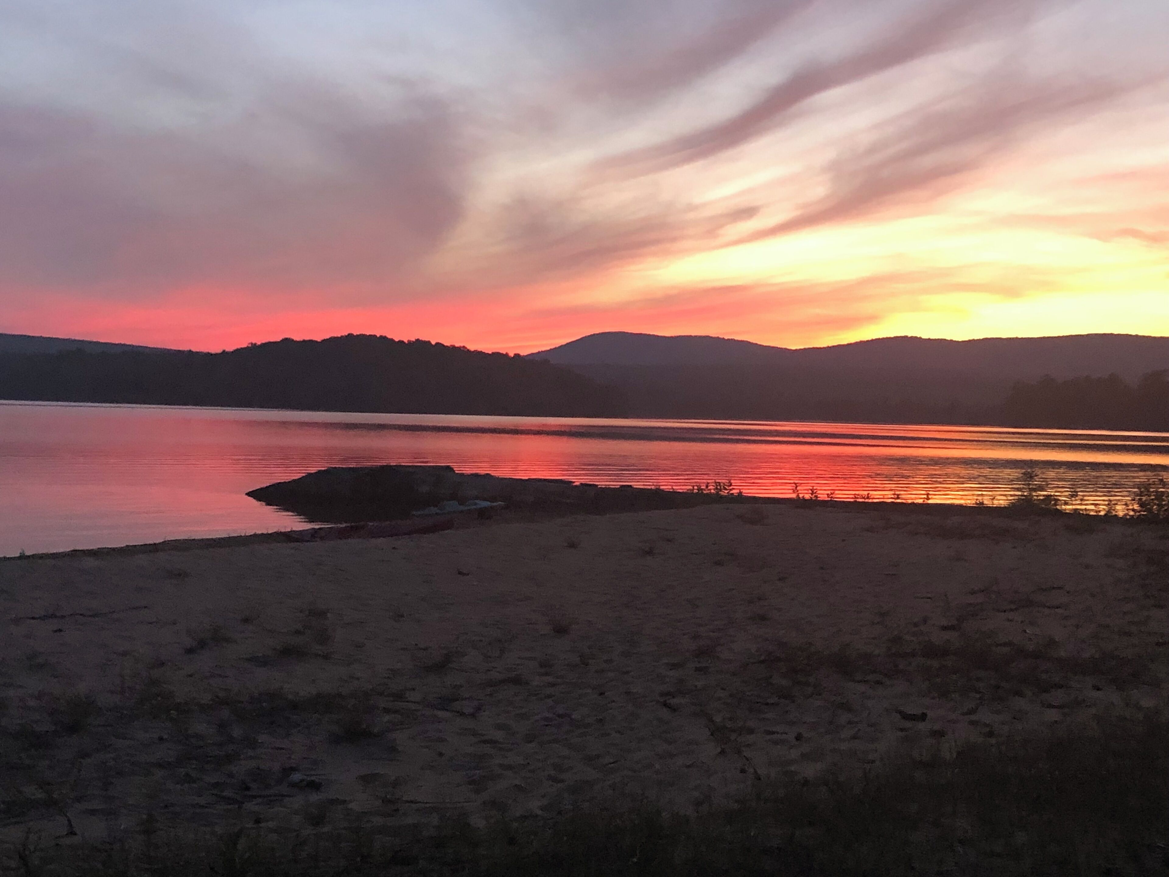 Un coucher de soleil sur un lac, avec une plage de sable et des collines à l’horizon.